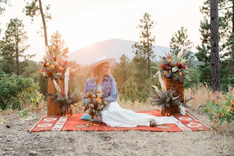 A woman reclines on a blanket between two floral arrangements on a vista holding a bouquet and wearing a hat.