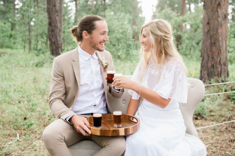 A groom and a bride cheers with mini beers and smile at each other. They're sitting in the forest.