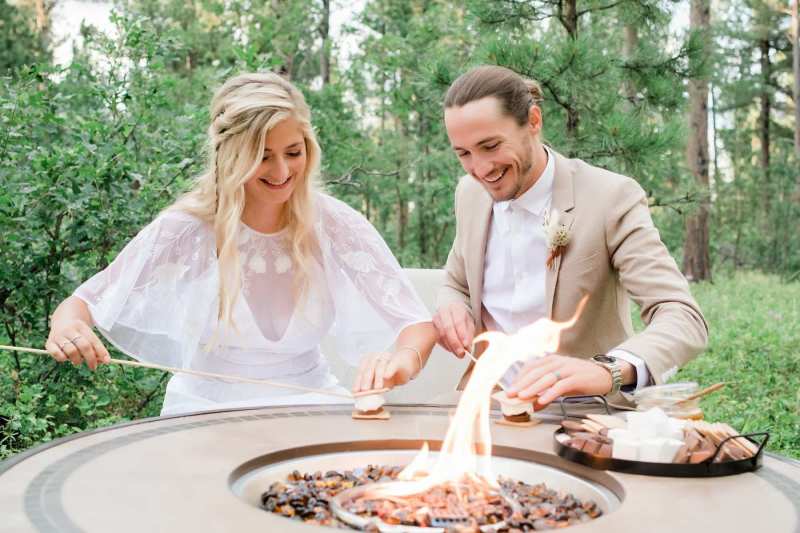 A bride and groom stand at a small fire preparing to make s'mores.