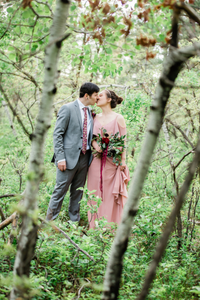 A groom in grey and a bride in mauve stand in the forest and kiss.