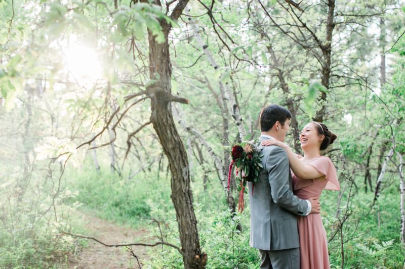 A groom in grey and a bride in mauve embrace and laugh. They're standing in the forest.