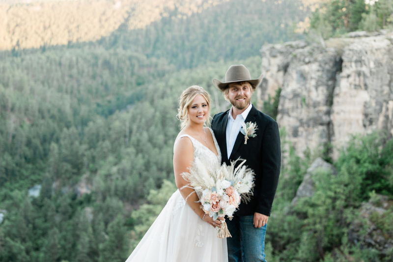 A bride in white holds a light colored bouquet as she stands with her groom wearing a cowboy hat. They're standing on a vista with mountains and evergreen trees in the background.