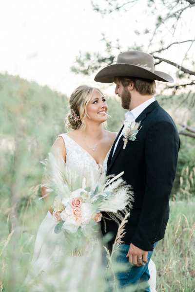 A bride and groom look at each other lovingly. She holds a bouquet and he wears a cowboy hat.
