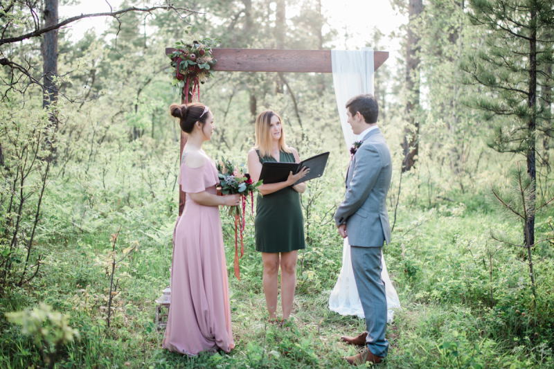 A bride in mauve and a groom in grey stand under an arbor draped in white as the female officiant  speaks.