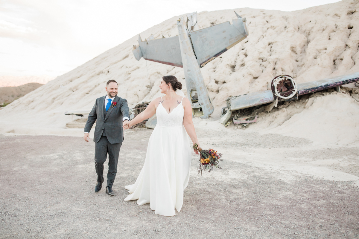 Bride and groom holding hands walking through the desert with airplane parts in the background.