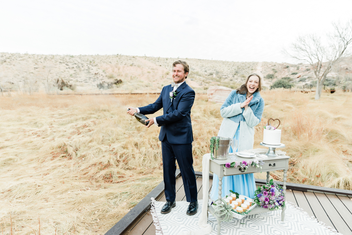 Groom popping bottle of champagne with bride next to him.