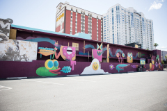 The side of a building is painted purple with a collection of strange and playful cartoon creatures lined up and looking out with smiles.