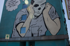 A mural of a retro-comic styled man with a skeletal face is sitting with a drink with a thought bubble coming from his head that says, “I gave her my heart, she left me for…”