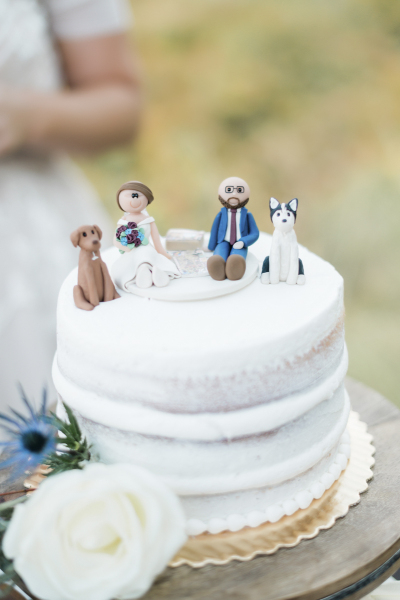 A cartoonish group of clay figurines depicting a sitting bride and groom and their dogs are used as a wedding cake topper.
