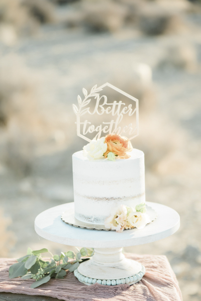 A small round wedding cake decorated with orange and white roses, a eucalyptus branch and a die cut cake topper which reads "Better together".