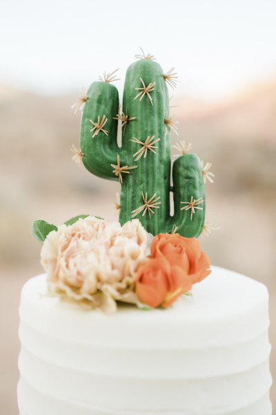 Close up photo of a wedding cake topped with a saguaro cactus statuette and pink and red decorative flowers.