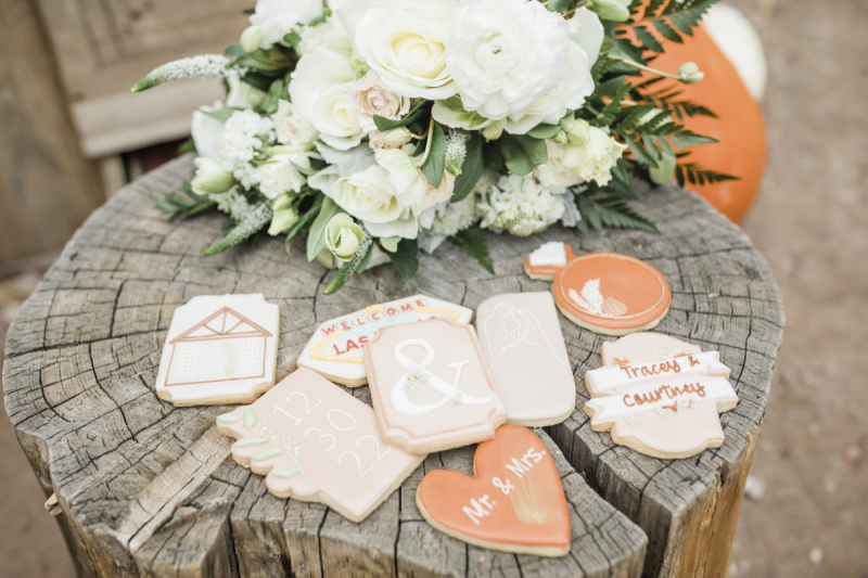 Personalized frosted sugar cookies are displayed with a bouquet of wedding flowers on top of a weathered log which acts as a table.