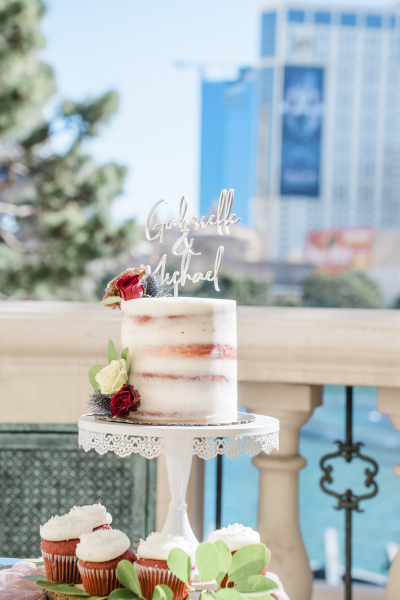 A red and white wedding cake sits on a lacey cake stand with cupcakes and a topper that reads "Gabrielle & Michael" in cursive lettering.
