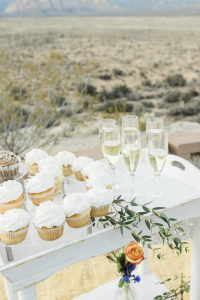 A white wooden serving cart filled with vanilla frosted cupcakes, flowers and glasses of champagne sits in a desert landscape.