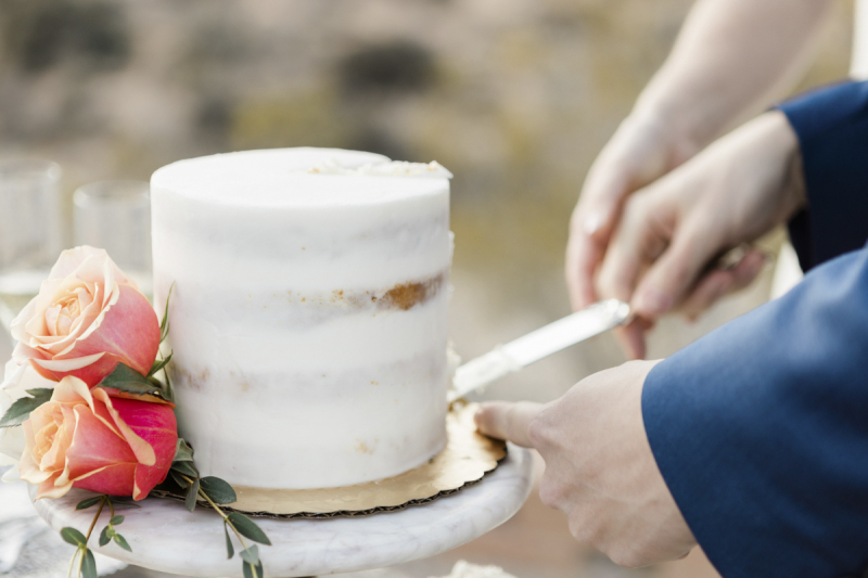Close up photo of a small round wedding cake being cut with a silver knife held by a couple's intertwined hands.