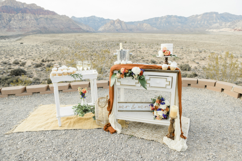 A pair of white wooden serving tables sitting on decorative rugs are displayed with cupcakes, champagne, wedding cake and flowers at the Red Rock Canyon overlook.