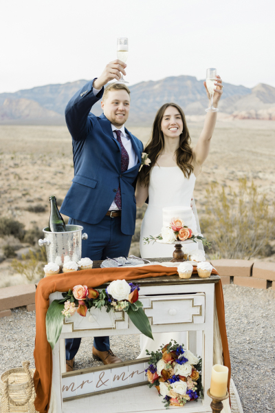 A smiling bride and groom stand behind a serving table stocked with champagne, flowers and cake as they raise their drinks high above their heads in salutation during their outdoor wedding with mountain views.