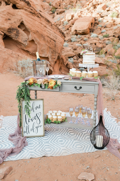 Wedding reception treat table display with cake, flowers, cupcakes and champagne arranged in front of a red sandstone outcropping.