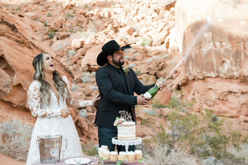 A bride with long blond hair and a groom wearing a black cowboy hat enjoy their desert southwest wedding ceremony celebration by spraying champagne from a bottle in front of a red sandstone outcropping.