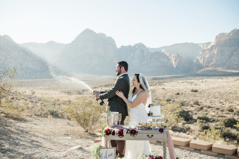 A groom sprays champagne out of a bottle while the smiling bride watches.