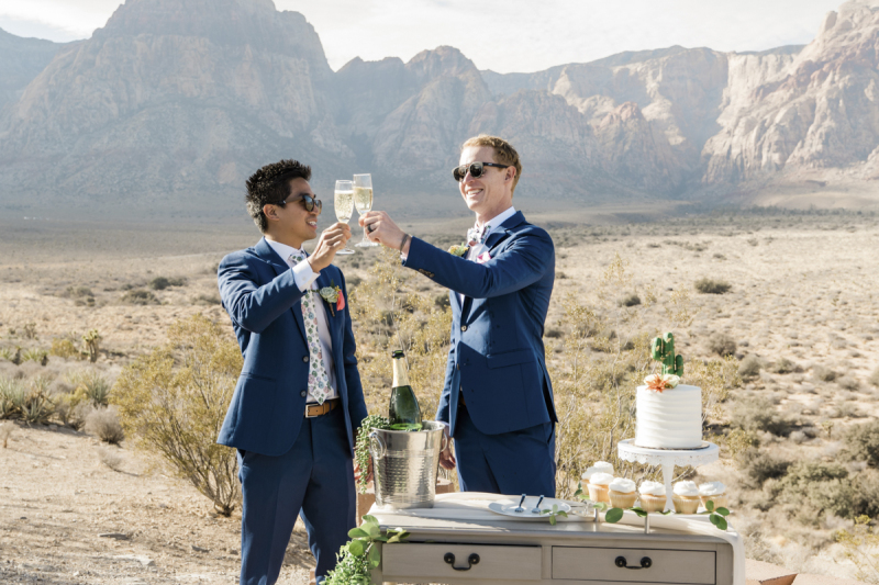 Two grooms toast each other with champagne flutes while wearing sunglasses and blue suits during their wedding ceremony celebration at Red Rock Canyon near Las Vegas.