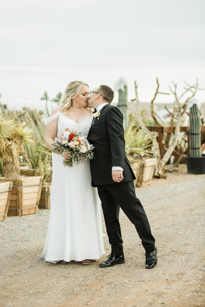 A bride receives a kiss from her groom as they pose for wedding photos in a cactus nursery.