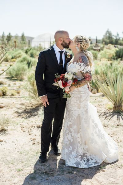 A bride and groom kiss in a desert landscape full of cacti and bushes.