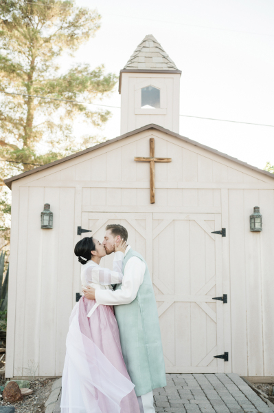 A bride and groom kiss in front of the doors of a small wooden chapel.
