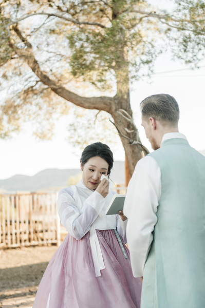 A bride wipes away tears as she reads her vows to her groom. There is a pine tree in the background.