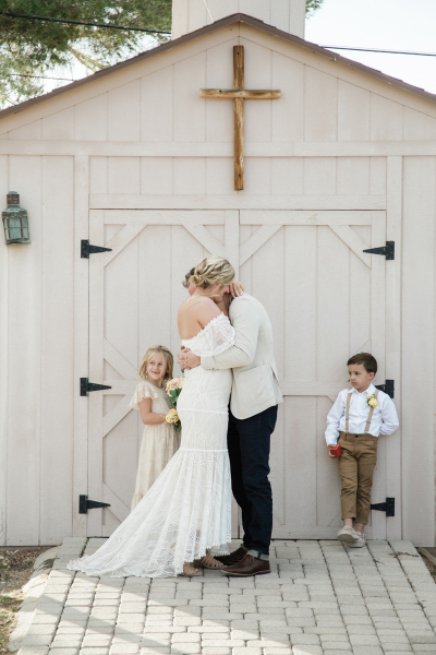 A bride and groom hug while two children stand beside them in front of a small wooden chapel. A bride and groom hug while two children stand beside them in front of a small wooden chapel.