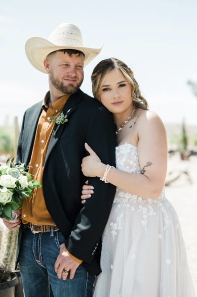 A bride holds tight to her groom's arm as they pose for wedding photos outside on a sunny day.