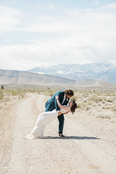 A groom dips his bride backwards on a dirt road with the snow-covered mountains of Red Rock Canyon in the background. A groom dips his bride backwards on a dirt road with the snow-covered mountains of Red Rock Canyon in the background.