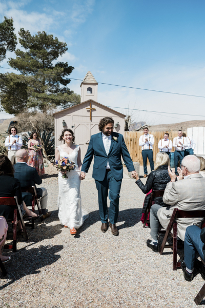 A couple walks towards the camera down an aisle leading away from a small garden chapel as wedding guests applaud