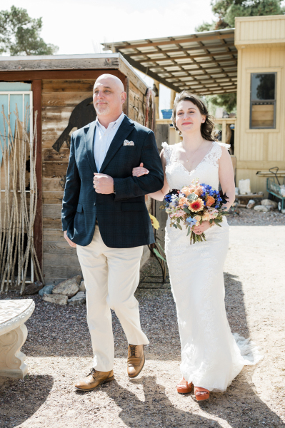 A smiling bride is escorted by a man past some wooden structures at Cactus Joe’s Blue Diamond Nursery. A smiling bride is escorted by a man past some wooden structures at Cactus Joe’s Blue Diamond Nursery.