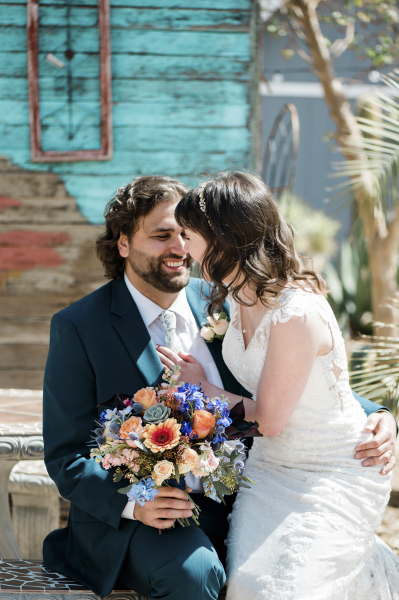 A groom and bride sit on a desert garden bench together and appear to be laughing.