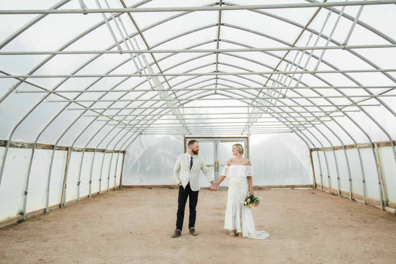 A groom and bride hold hands while standing in the center of an empty greenhouse. A groom and bride hold hands while standing in the center of an empty greenhouse.