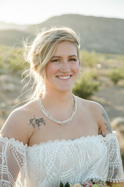 A portrait of a bride wearing a lacy shoulder-less dress standing in the desert.