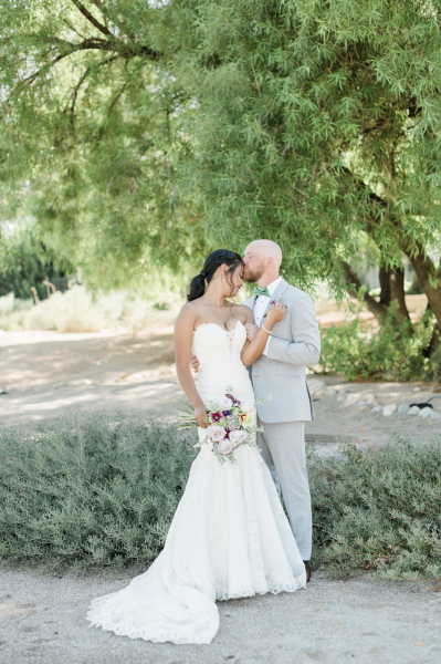 Groom kissing bride's forehead in front of trees.