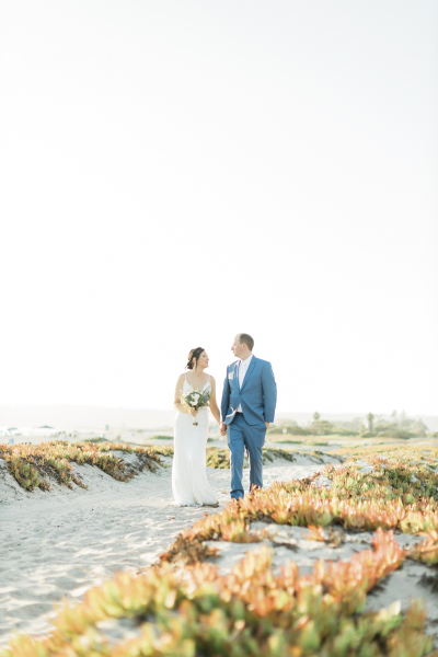 Newlywed couple walking hand in hand on Coronado Beach.