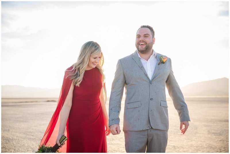 A woman in a flowy bright red dress and a man in a gray suit walk together and laugh. They are celebrating their wedding on a sunny day at the Dry Lake Bed in Boulder City, Nevada.