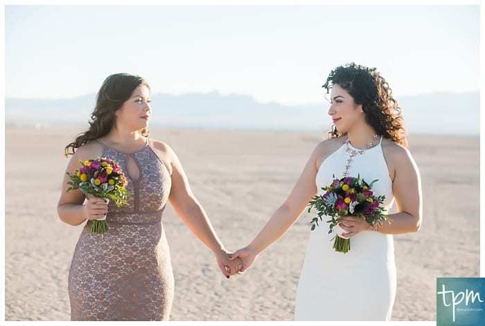 Two women hold hands and stand together on the Dry Lake Bed south of Boulder City near Las Vegas, Nevada. They are each holding a small bouquet of flowers. They are both wearing elegant sleeveless dresses.