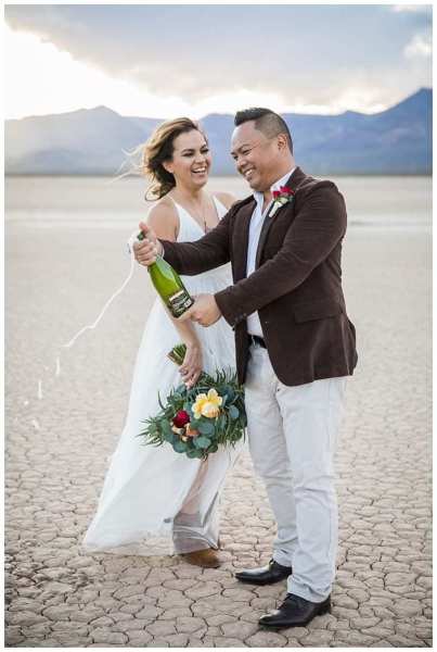 A bride and groom smile and laugh as he pops the cork on a bottle of sparkling wine. They are celebrating their wedding at the Dry Lake Bed in Boulder City, Nevada near Las Vegas.