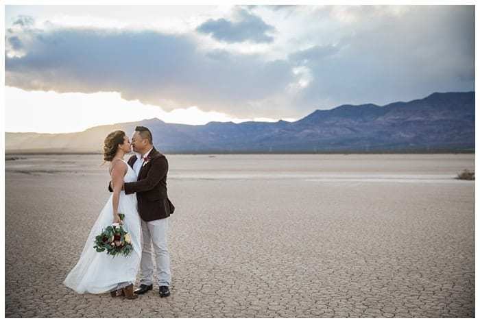 Moody clouds move in over the Eldorado Dry Lake Bed as a bride and groom embrace while posing for wedding day photos. The wide shot shows the cracked and dried out ground, and the mountains rising in the distance.
