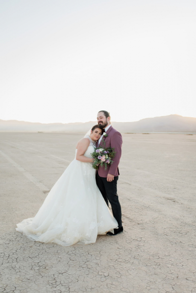 The sun sets behind the mountains at the Dry Lake Bed near Las Vegas, Nevada while a bride leans on her groom's chest. They are posing for photos on their wedding day.