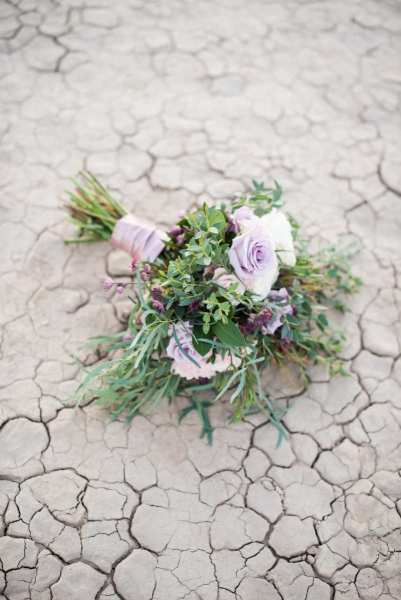 A wedding floral bouquet sits on the parched ground of the Eldorado Dry Lake Bed outside of Las Vegas, Nevada. The purple and pink flowers are wrapped in a ribbon and accented by greenery.