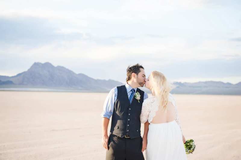 A groom kisses his bride with a lacy white backless wedding dress. They have gotten married on the Dry Lake bed south of Boulder City, Nevada. Mountains are seen in the background.