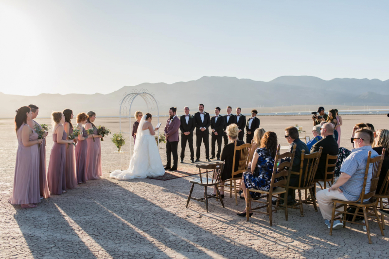 A wedding is being held at the Dry Lake Bed in Las Vegas. At the center is the bride and groom and the officiant. They are flanked by their bridesmaids and groomsmen. Several guests sit in chairs arranged in front of them.