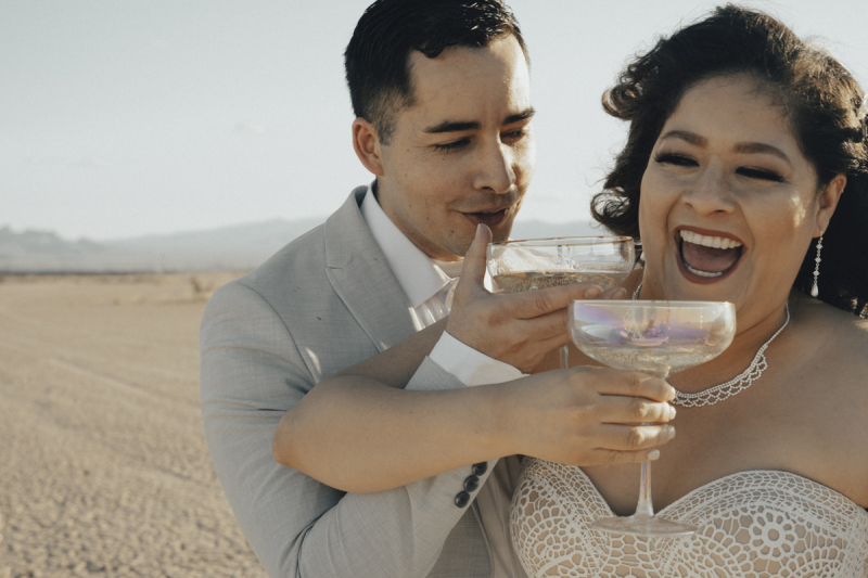 A bride and groom lock arms and drink Champagne from coupe glasses. They are toasting their marriage following their wedding on the Eldorado Dry Lake Bed outside of Boulder City and Las Vegas, Nevada. A bride and groom lock arms and drink Champagne from coupe glasses. They are toasting their marriage following their wedding on the Eldorado Dry Lake Bed outside of Boulder City and Las Vegas, Nevada.