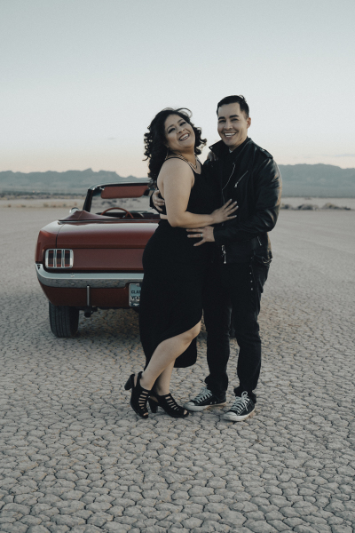 A man and woman both wearing black hold each other and smile as they stand in front of the rear end of a red 1965 Ford Mustang convertible that is parked on the Eldorado Dry Lake Bed.
