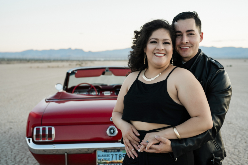 A close up photo of a man and woman holding each other and smiling as they stand in front of the rear end of a red 1965 Ford Mustang convertible that is parked on the Eldorado Dry Lake Bed.